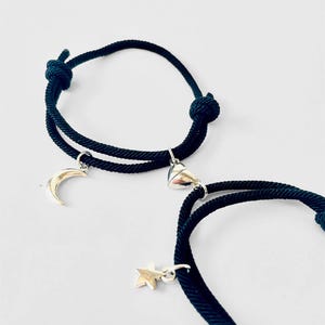 May include: Two black string bracelets with silver charms. One bracelet has a crescent moon charm and the other has a heart charm.