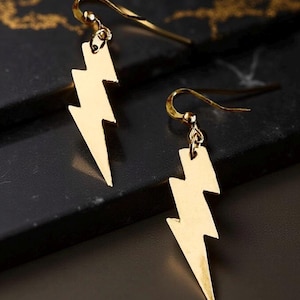 May include: A pair of gold-colored lightning bolt earrings. Each earring features a stylized lightning bolt pendant suspended from a curved ear wire. The earrings are displayed against a dark background.