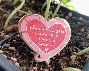 May include: A heart-shaped enamel pin with the text "Would You Still Love me if I was a worm?" in white lettering. The pin has a pink border and a red center, and a small worm illustration.