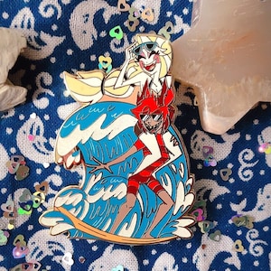 May include: Gold-tone enamel pin featuring a red and white cartoon character riding a surfboard with a blonde cartoon character on top of the wave. The pin is on a blue and white fabric background with a white star-shaped object in the upper right corner.
