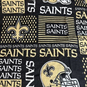 May include: Black and gold fabric with a repeated pattern of the New Orleans Saints logo, including a football helmet and fleur-de-lis. The text "Saints" is repeated throughout the fabric.