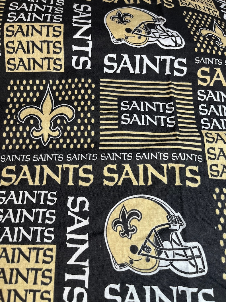 May include: Black and gold fabric with a repeated pattern of the New Orleans Saints logo, including a football helmet and fleur-de-lis. The text "Saints" is repeated throughout the fabric.