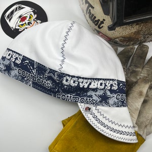 May include: White welding cap with a blue and white Cowboys print. The cap has a zig-zag stitch around the edge.
