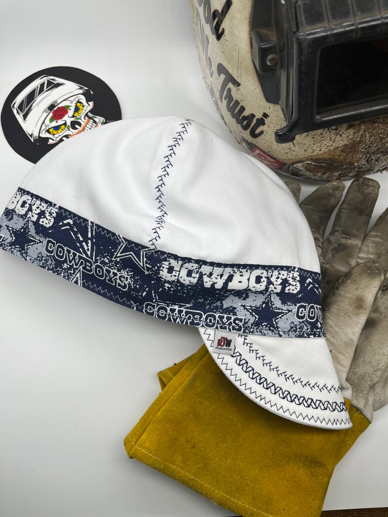 May include: White welding cap with a blue and white Cowboys print. The cap has a zig-zag stitch around the edge.