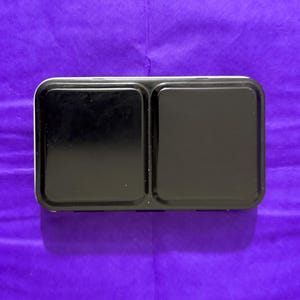 Puede incluir: Una paleta rectangular de pintura de acuarela negra con dos pozos grandes y vacíos. La paleta tiene un borde blanco y está sobre un fondo morado. Ideal para artistas y pintores.