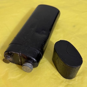 Könnte beinhalten: Eine schwarze, rechteckige Flasche mit abgerundeter Oberseite und einem separaten, passenden Deckel. Die Flasche hat einen Metallverschluss und ein kleines, rundes Metallstück an der Seite. Die Flasche liegt auf einer gelben Oberfläche.