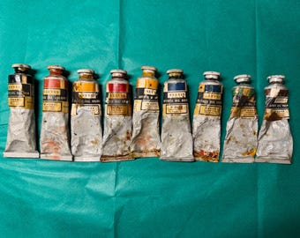 May include: A collection of nine used tubes of Reeves Artists' Oil Colour paint, each with a different color. The tubes are silver with colorful caps and labels, arranged on a teal green surface. The tubes are partially squeezed.