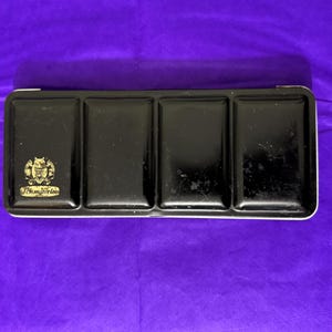 以下が含まれることがあります： 4つのウェルを備えた黒い長方形の水彩絵の具パレット。パレットには金のエンブレムと「Schmincke」の文字が前面に印刷されています。パレットは紫色の背景にあります。