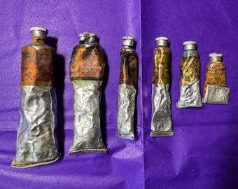 May include: A collection of six vintage paint tubes, each with a metallic silver body and a copper-colored top. The tubes are partially squeezed, showing signs of use. The background is a solid purple color.
