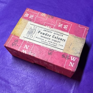 May include: A vintage box of Winsor & Newton Powder Colours in Winsor Violet. The box is a faded pink with a beige label and text that reads "Artists' Quality". The box is on a purple surface.