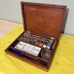 Puede incluir: Caja de pintura de acuarela de madera abierta con una variedad de cuadrados de pintura coloridos. La caja contiene una paleta blanca, un vaso pequeño y una borla azul. La caja está abierta, revelando las pinturas y accesorios.