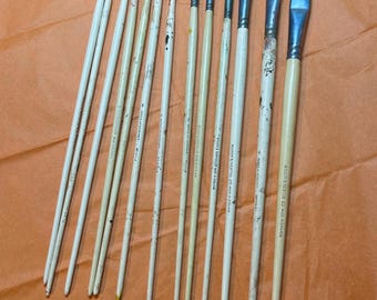 May include: A collection of paint brushes with wooden handles and metal ferrules. The brushes vary in size and bristle shape, with some showing signs of use. The bristles are in various colors, including white, brown, and green. The brushes are arranged on an orange surface.