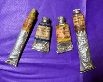 May include: Four vintage oil paint tubes on a purple surface. The tubes are labeled with names like "LECHRON YELLOW" and "COBALT BLUE TINT." The tubes are silver with labels in English. The paint tubes are from "REEVES SCHOOL QUALITY."