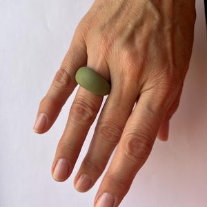 Puede incluir: Un anillo verde oliva liso. El anillo tiene una forma redondeada y se lleva en un dedo. El anillo es de diseño sencillo y moderno. El anillo está hecho de un material sólido.