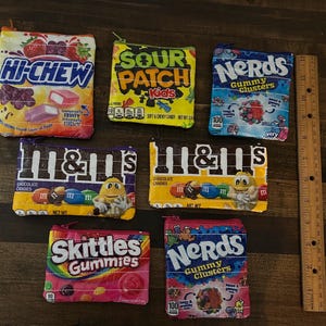 May include: A collection of colorful zippered pouches featuring candy brands like Hi-Chew, Sour Patch Kids, Nerds, M&M's, and Skittles. The pouches are rectangular and have various colors and graphics associated with each candy.
