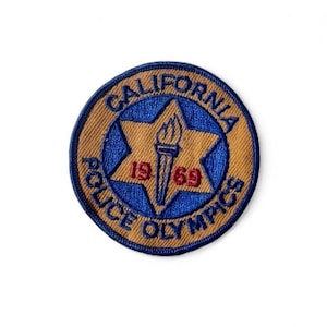 May include: A circular patch with the words "CALIFORNIA POLICE OLYMPICS" in blue lettering. A blue star with a torch in the center and the numbers "19 69" in red. The patch has a tan border.