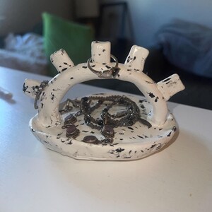 Op de afbeelding: Een witte keramische sieradenhouder met zwarte spikkels. De houder heeft een cirkelvormige basis en een gewelfde bovenkant met verschillende palen. Ringen en kettingen worden op de houder getoond.