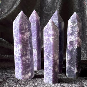 May include: Six purple lepidolite crystal points, each with a unique pattern of white and purple swirls. The points are arranged in a row on a dark gray surface.