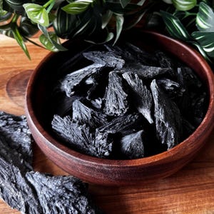 May include: A wooden bowl filled with black kyanite crystals. The crystals have a spiky, radiating structure and a dark, charcoal color. Green leaves are visible in the background, adding a natural element to the composition.