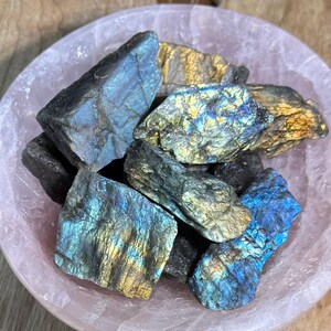 May include: A collection of raw labradorite stones in a light pink bowl. The stones display iridescent flashes of blue, gold, and green. The bowl is round and has a smooth surface. The stones vary in size and shape, with rough, textured surfaces.