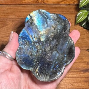 May include: A polished labradorite stone with a unique, organic shape. The stone displays iridescent flashes of blue, gold, and green across a dark gray base. The surface has a textured appearance, and the stone is held in a hand.