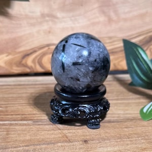 May include: A clear, round crystal ball with black inclusions sits on a black, ornate stand. The crystal has a gray and black color scheme. The stand has a detailed, carved design. The background is a wooden surface.