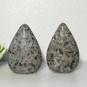May include: Two gray teardrop-shaped decorative stones with black speckles. The stones have a polished surface and a subtle, carved design near the top. They are displayed on a white surface.