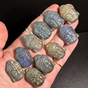 Könnte beinhalten: Eine Sammlung kleiner, geschnitzter Buddha-Kopf-Figuren in verschiedenen Grau-, Blau- und Goldtönen. Die Figuren bestehen aus einem steinartigen Material und werden in einer Hand gehalten. Die Buddha-Köpfe weisen detaillierte Gesichtszüge auf.
