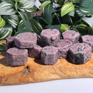 以下が含まれることがあります： さまざまなサイズの、立方体に近い形状のラフなルビーの結晶のコレクション。結晶は濃いピンクがかった赤色で、濃い緑色のインクルージョンがあります。天然の木製の表面に置かれ、背景には緑の葉があります。