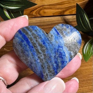 May include: A heart-shaped stone with a striking blue and gray pattern. The polished surface of the stone reveals a mix of deep blue and gray hues, creating a marbled effect. The heart is held in a hand, showcasing its size and shape.