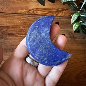 May include: A crescent moon-shaped lapis lazuli stone. The stone is a deep blue color with flecks of gold throughout. The moon is held in a hand, with a wooden surface and greenery in the background.