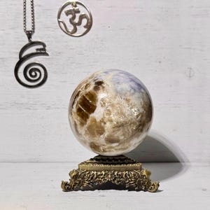 May include: A large, round, polished, light brown and white gemstone sphere on a gold metal stand with ornate details.