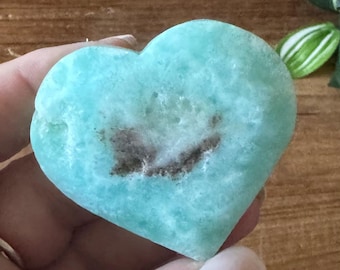 Peut inclure: Une pierre précieuse en forme de cœur, bleu clair, avec un centre marbré brun. La pierre a une surface rugueuse et texturée et est tenue dans une main. L'arrière-plan est une surface en bois floue avec un soupçon de verdure.