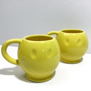 May include: Two yellow ceramic mugs with a smiley face design. Each mug has a rounded shape, a handle, and a base. The mugs are a bright yellow color and are suitable for coffee, tea, or other beverages.