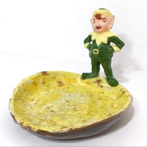 May include: A vintage ceramic ashtray featuring a green-clad elf figurine with a yellow collar and cap. The ashtray has a yellow and brown speckled interior and a brown exterior.