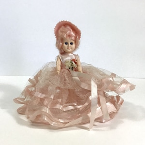 May include: A vintage pink plastic doll with pink hair, wearing a pink dress and a pink hat. The doll is holding a bouquet of flowers and is sitting on a pink tulle skirt.