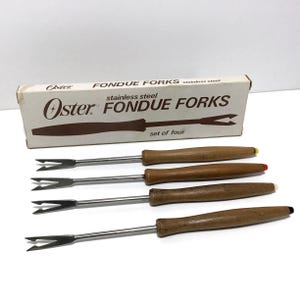 May include: Set of four vintage Oster stainless steel fondue forks with wooden handles. The forks have a two-pronged design and are displayed with their original box. The box is white with brown text and the Oster logo.