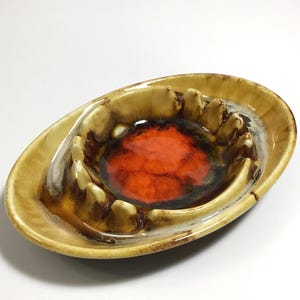 May include: Oval-shaped ceramic ashtray with a brown and tan exterior and a vibrant orange interior. The ashtray features a raised rim with multiple resting points for cigarettes. A vintage decorative piece.