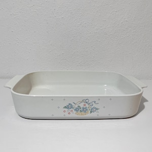 May include: A rectangular white ceramic baking dish with integrated side handles. The exterior features a central floral motif depicting a basket filled with blue and pink flowers, accented by a light blue ribbon and small scattered diamond patterns.