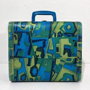 May include: A rectangular suitcase featuring a vibrant geometric pattern in shades of blue, light green, and dark green. The case has a solid blue plastic handle attached to the top center and is constructed with visible stitching along the edges.