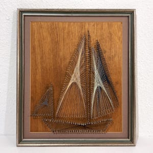 May include: A framed string art piece depicting a sailboat on a wood grain background. The artwork uses silver nails and thin threads in shades of cream, blue, and brown to form the hull and sails of the boat, enclosed in a metallic silver frame.