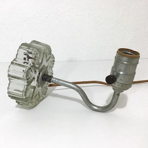 May include: An antique clear glass lamp with a starburst design. The lamp has a metal arm and a bulb socket. The lamp is on a white surface.