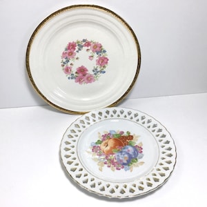 May include: Two decorative porcelain plates. The larger plate has a gold rim and a floral design with pink and blue flowers. The smaller plate has a white, openwork border and a fruit design with an orange peach and purple plums.
