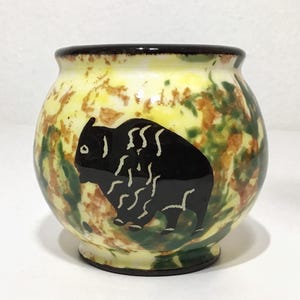 May include: A small, round ceramic pot with a black bison design. The pot has a yellow, green, and brown mottled background. The rim is dark brown. The bison is black with white detailing.