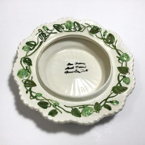 May include: Off-white ceramic dish with a scalloped edge and a central recessed area. The rim is decorated with green vines and leaves. The bottom of the dish has text that reads "Pottery Hand Painted Greenville, J.A."