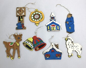 May include: Nine wooden Christmas ornaments, including a reindeer, a clock, a church, a bell, a house, a lamb, a monk, and two ornaments with geometric designs. The ornaments are painted in various colors, including red, blue, green, yellow, and white.