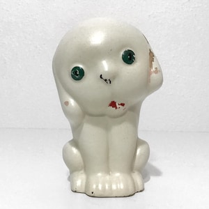 May include: A vintage white ceramic dog figurine with green eyes and red accents on the mouth. The dog is sitting upright with its head tilted slightly. The figurine has a glossy finish and shows signs of wear.