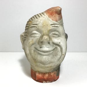 May include: Vintage, hand-painted ceramic head with a smiling face, wearing a red bow tie and a cap with a red accent. The head has stitched details and is painted in shades of gray, white, and red. A unique, antique decorative piece.