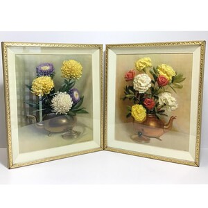 May include: Two framed floral still life art pieces. One features yellow, white, and purple flowers in a silver teapot. The other displays red, white, and yellow flowers in a copper teapot. Both are set against a neutral background.