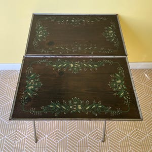 May include: A rectangular folding table with a dark brown wood grain surface featuring a decorative green and cream floral border pattern. The table has a thin metallic silver frame and slender metal legs, providing a functional surface.